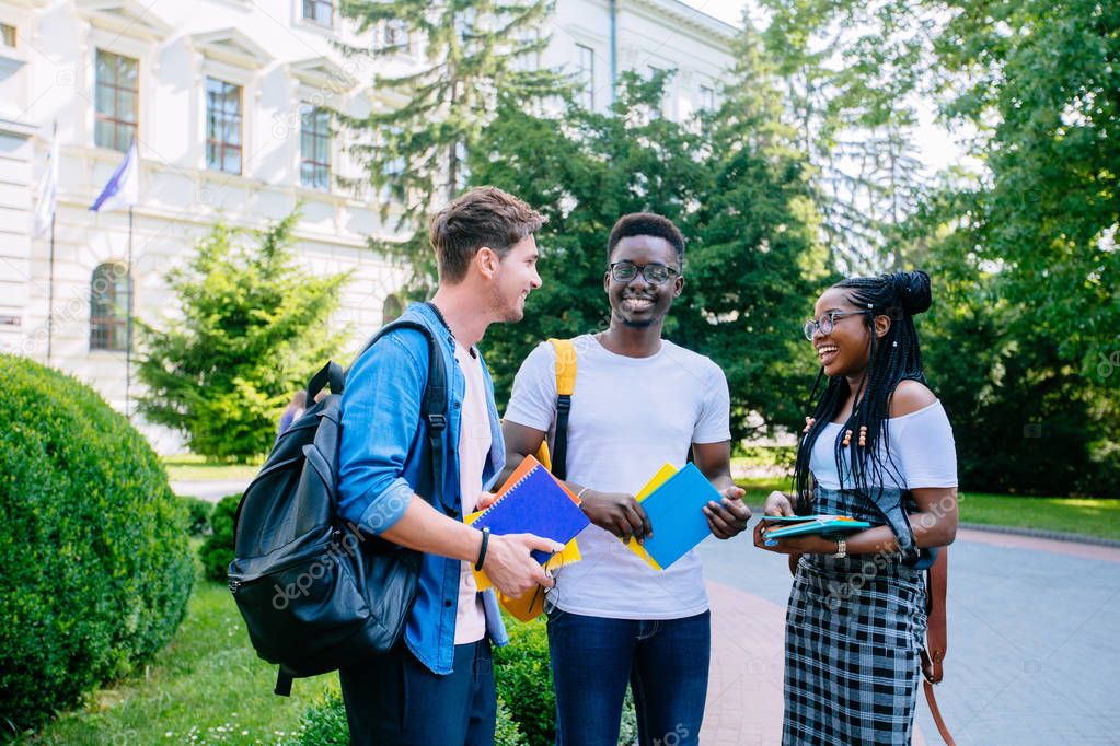 Laughing african american female student with backpack with two males multiethnic friends talking outdoor over university buildig.