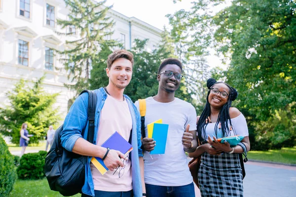Portrait de trois lycéens multiethniques heureux de différents pays marchant vers la classe, après la leçon, en plein air tenant des livres dans la ville européenne. Concept d'amitié multinationale . — Photo