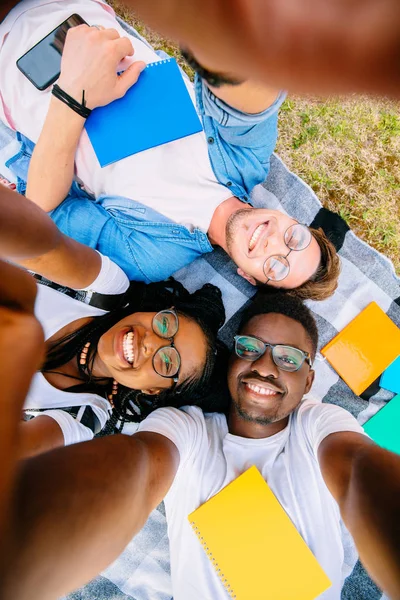 Young handsome beard student man taking self-portrait to beautiful black girlfriend with her african brother. Top view. Multiethnic friends lying on grass outdoor.
