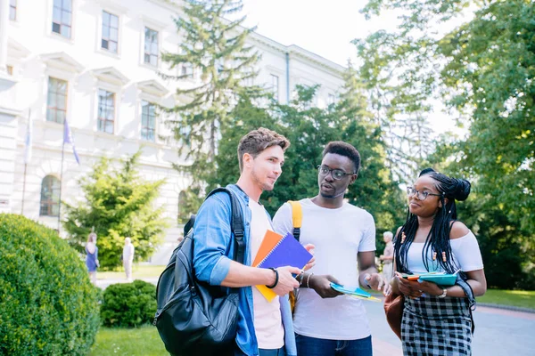 Trois étudiants positifs de différents pays avec des livres et des sacs à dos parler tout en marchant contre la construction dans le campus universitaire en plein air. Nouveau concept d'année académique . — Photo