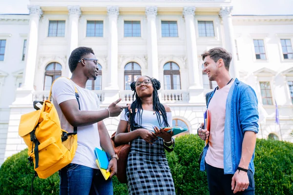 Trois étudiants positifs de différents pays avec des livres et des sacs à dos parler tout en marchant contre la construction sur le fond dans le campus universitaire en plein air. Nouveau concept d'année académique . — Photo