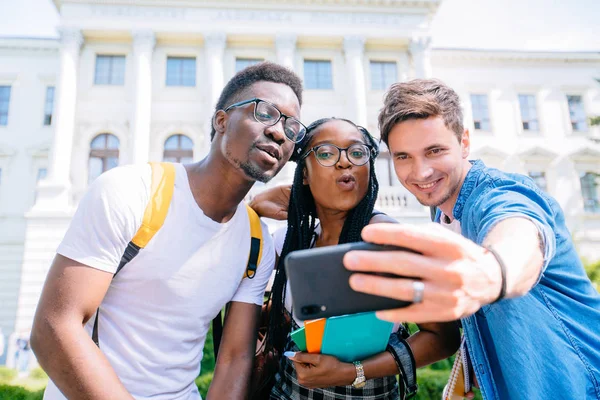 Photo de groupe multiethnique de trois jeunes étudiants heureux debout à l'extérieur font selfie par téléphone. En regardant de côté . — Photo