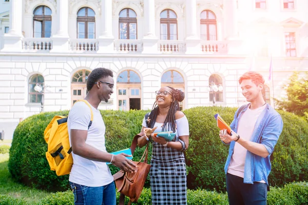 Tres estudiantes positivos de diferentes países con libros y mochilas hablando mientras caminan contra la construcción en el campus universitario al aire libre. Nuevo concepto de curso académico . — Foto de Stock