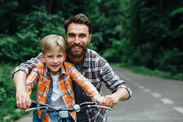 Портрет улыбающегося отца и его сына, веселящихся вместе в зеленом парке, обучающих сына кататься на велосипеде. Концепция досуга . — стоковое фото