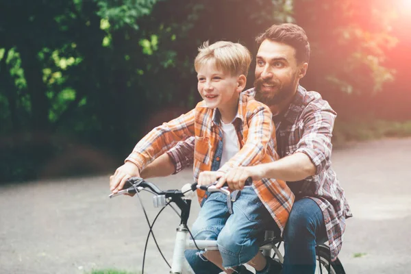 Отец и сын веселятся вместе, катаясь на велосипеде, прогуливаясь на рейде по зеленому парку или лесу. Папа показывает руками вперед, оба смеются. Тонко. Концепция досуга . — стоковое фото