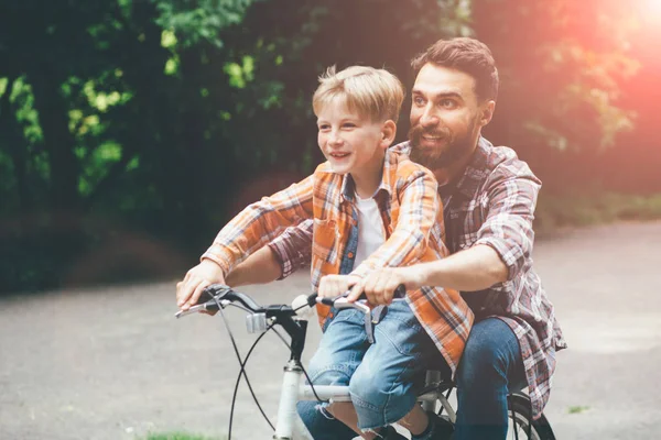 Father and son having fun together riding bicycle together, walking on raod in green park or forest. Daddy pointing by hands forward, both laughing. Toned. Adventure leisure concept.