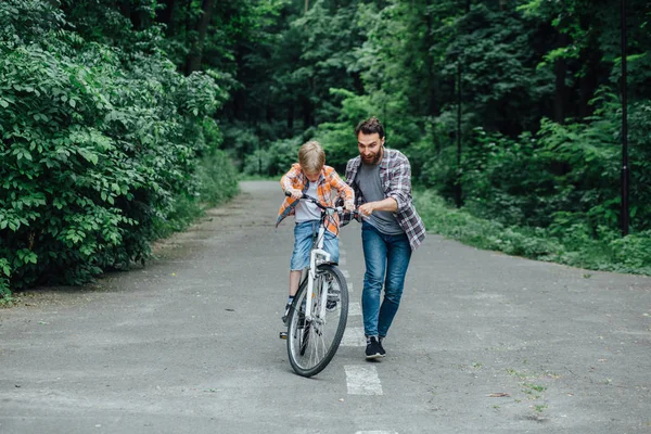 Feliz padre risueño con su hijo en una bicicleta de equilibrio, familia divirtiéndose juntos en el parque verde, papá enseñando a su hijo a montar en bicicleta. Aventura concepto de ocio . —  Fotos de Stock