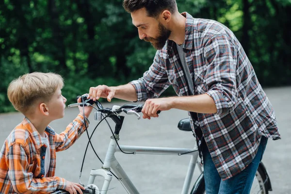 Красивый молодой папа и его милый маленький велосипед в зеленом летнем парке. Отец осматривает велосипед сына. . — стоковое фото