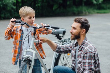 Yakışıklı baba ve parkta bisiklet ile onun sevimli küçük oğlu, birbirlerine bakıyor ve gülümsüyor. Baba oğlunun bisikletini inceliyor..