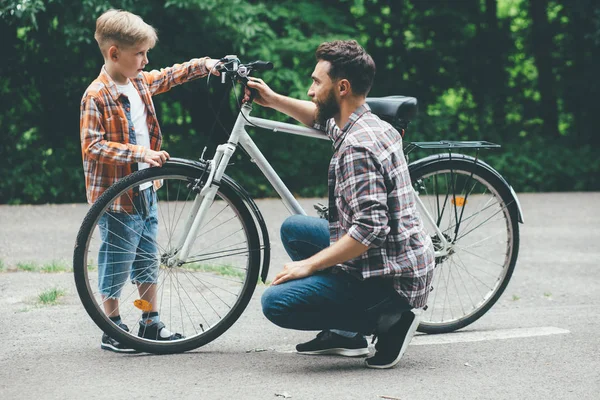 Бородатый кавказский отец и мальчик смотрят на старый неисправный велосипед. Отец и его сын ремонтируют велосипеды на улице в зеленом парке на дороге . — стоковое фото