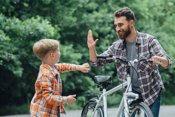 父は息子のために古い自転車をあげる。公園でサイクリングしながらハイ5を与える幸せな子供の男の子. — ストック写真