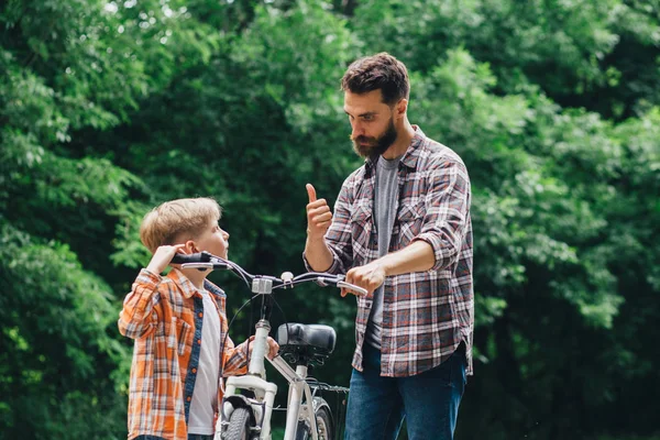 Handsome beard father talking with his son riding a bicycle and showing thumb up at summer park green leaves on background. — Stock Photo, Image