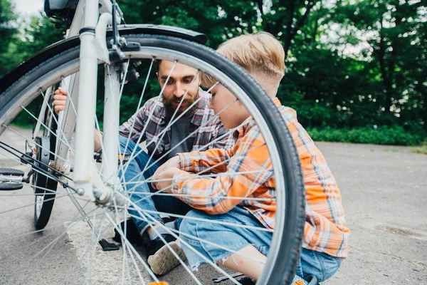 Взрослый хипстерский бородатый отец и его младший сын в клетчатой рубашке вместе ремонтируют велосипед в солнечный летний день в парке . — стоковое фото