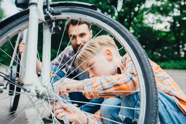 An adult hipster beard father and his elementary age little son wearing in plaid shirt repairing bicycle together outside on a sunny summer day in park.