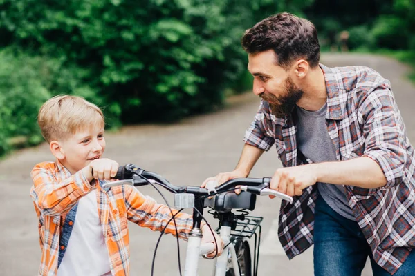 Счастливая семейная концепция. Красивый папа и его милый маленький сын с велосипедом в парке, смотрят друг на друга и улыбаются. Отец осматривает велосипед сына. . — стоковое фото