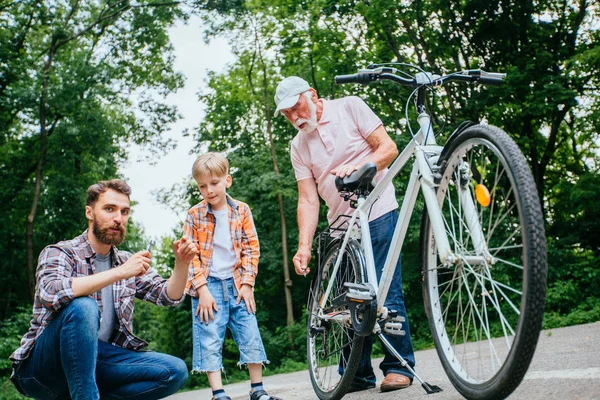 Отец учит сына ремонтировать велосипед на открытом воздухе. Папа, дедушка и внук школьного возраста ремонтируют велосипед в летнем зеленом парке. Счастливые отношения семьи концепция другого поколения . — стоковое фото