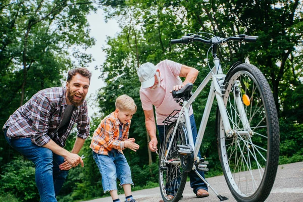 Смеющийся красивый отец, дедушка и сын школьного возраста ремонтируют старый велосипед на открытом воздухе в летнем парке. Разное поколение проводит время вместе концепция . — стоковое фото