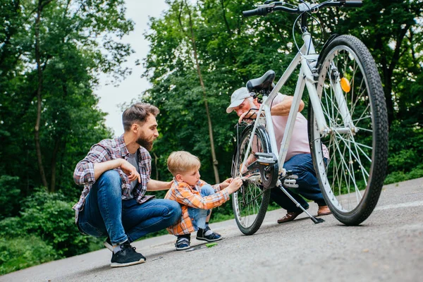 父は息子に屋外で自転車を取り返し教えている。お父さん、おじいちゃんと学校時代の孫の息子は、外の夏の緑の公園で自転車を修理します。幸せな家族関係異なる世代の概念. — ストック写真