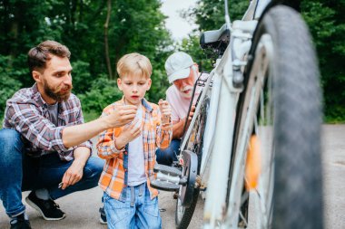 Baba açık havada bisiklet reparing için araç oğlu veriyor. Baba, büyükbaba ve okul çağındaki torunun oğlu dışarıdaki yazlık parkta bisiklet tamir ediyor. Aile ilişkisi farklı nesil kavramı.