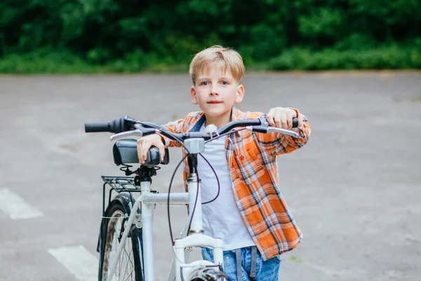 Мальчик в оранжевой рубашке носит большой велосипед в летнем парке под открытым небом . — стоковое фото