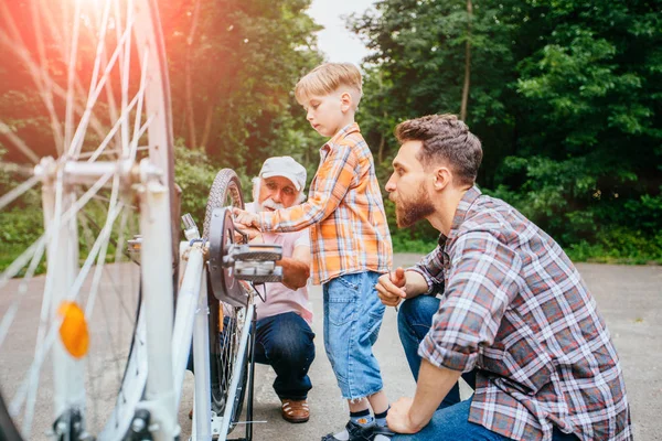 Senior man helping his son and grandson fixing upside down bike outdoor in city park in summer day. Family relation different generation concept.