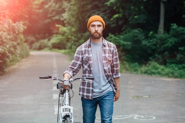 Hadsome hipster man carries a bike outdoor Healthy lifestyle concept. Hansome male in street wear, walking with his bike beside him holding it by the handlebar along road on sunny summer day