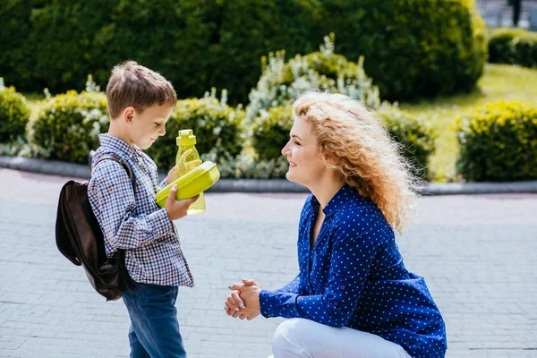 Blonde krullend Kaukasische moeder geeft school lunch en fles met wather naar haar kleine zoon buitenshuis in zonnige ochtend. Terug naar school concept. — Stockfoto