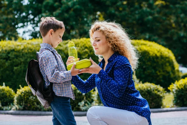 Gelukkig positieve Kaukasische moeder in blauw shirt geven school lunch en fles met wather aan haar kleine leerling zoon buitenshuis in zonnige ochtend. Terug naar school. onderwijs, zorgzaam, Eenouder concept. — Stockfoto