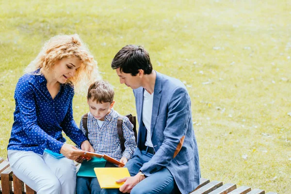 La madre rubia rizada y el padre están sentados en el banco y enseñando a un alumno en el parque con libro al aire libre. concepto de familia feliz juntos . — Foto de Stock