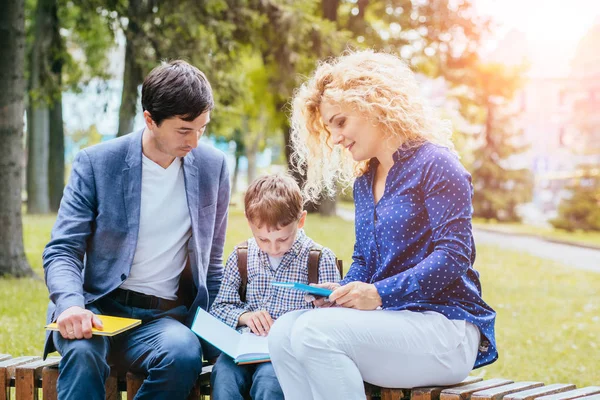 La madre rubia rizada y el padre están sentados en el banco y enseñando a un alumno en el parque con libro al aire libre. concepto de familia feliz juntos . — Foto de Stock