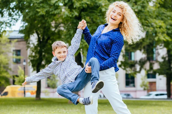 Blonde krullend moeder in blauw shirt en haar zoon genieten van Quality Time in Park, moeder het gooien van haar zoon, Kaukasische moeder en zoon concept. — Stockfoto