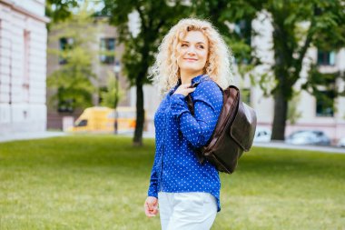 Mavi gömlek ve kahverengi deri bacpack, açık portre sevimli sarışın kıvırcık saçlı kadın.