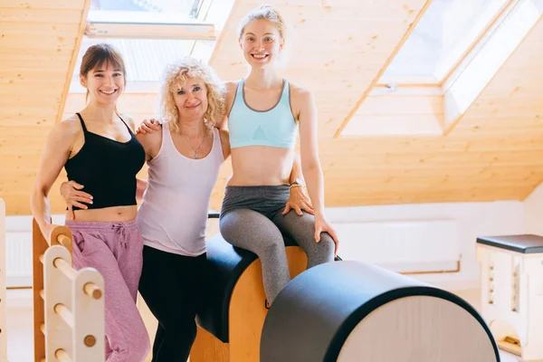 Portrait of three different age sporty women friends hugging, sitting on pilates barrel equpment ready to start workout together yoga or pilates exercise indoor. Sport friendship concept.