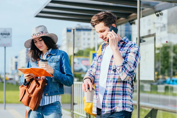 Estudiantes pareja viajero esperando tranvía en parada.. Guapo chico hablando de teléfono mientras que la mujer elegante lectura libro conspectus en la estación de autobuses con paisaje urbano . — Foto de Stock