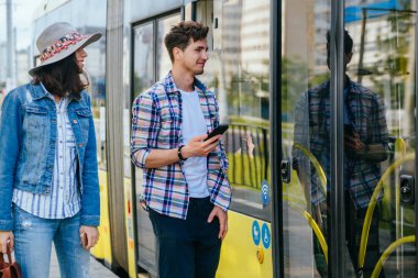 Genç kadın ve erkek yolcuların yaşam tarzı fotoğrafı tramvaykapısını açarken bekleyen telefonları tutuyor. Tramvay durağında yakışıklı adam ile konuşurken sırt çantası ile kadın gezgin Portresi.