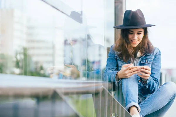 Stylish hipster girl chatting in network via smart phone with her friends while sitting on bus stop in summer day,fashionable woman tourist searching information about bus traffic using cell telephone