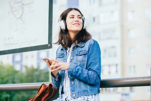 Sonriente mujer morena emocionada con chaqueta de mezclilla azul con auriculares mirando hacia otro lado, relajante, escuchando música mientras espera en la parada de tranvía en una ciudad . — Foto de Stock