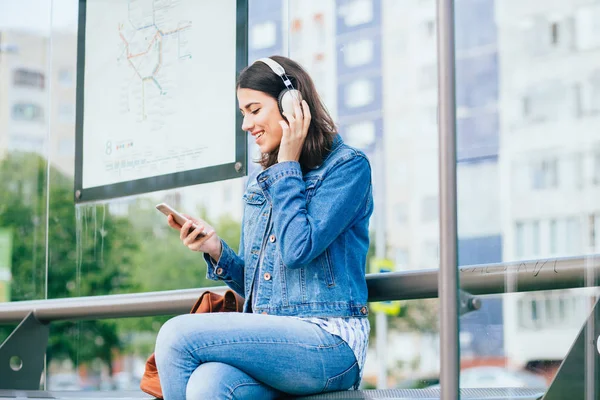 Sonriente mujer morena emocionada con chaqueta de mezclilla azul con auriculares mirando hacia otro lado, relajante, escuchando música mientras espera en la parada de tranvía en una ciudad . — Foto de Stock