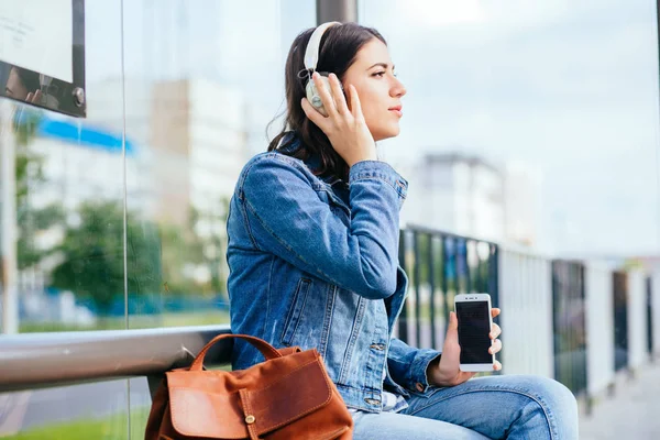 Sonriente mujer morena emocionada con chaqueta de mezclilla azul con auriculares mirando hacia otro lado, relajante, escuchando música mientras espera en la parada de tranvía en una ciudad . — Foto de Stock