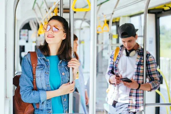 Joven adorable mujer alegre está de pie en el autobús usando el teléfono y sonriendo . — Foto de Stock