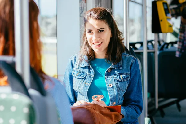 Duas jovens mulheres bonitas e alegres estão sentadas na frente umas das outras em um ônibus ou bonde e olhando para os livros, lendo, falando sorrindo enquanto esperam por um ônibus para levá-los ao seu destino . — Fotografia de Stock