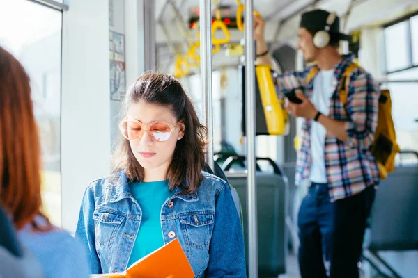 Dos guapas y alegres mujeres jóvenes están sentadas una frente a la otra en un autobús o tranvía y mirando los libros, leyendo, hablando sonriendo mientras esperan que un autobús las lleve a su destino. . — Foto de Stock