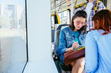 İki neşeli güzel genç kadın bir otobüs veya tramvay da birbirlerinin önünde oturuyor ve kitaplara bakarak, okuma, konuşurken genç yakışıklı adam arka planda müzik dinlerken.