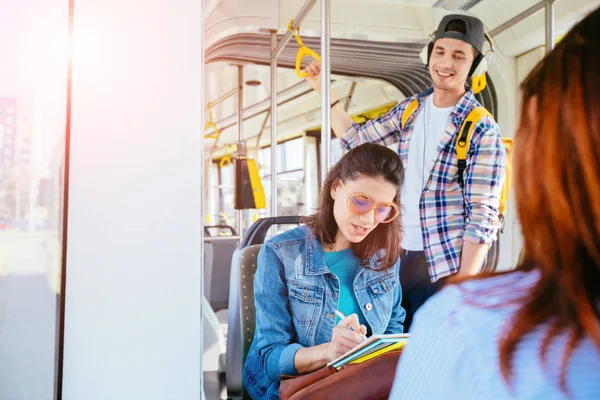 Dos mujeres jóvenes alegres y bonitas están sentadas una frente a la otra en un autobús o tranvía y mirando los libros, leyendo, hablando mientras un joven guapo escucha música de fondo . — Foto de Stock