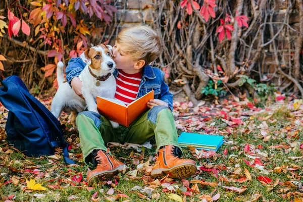 School boy sitting on bright colorful leaves kissing and hugging his dog in the fall autumn park, outdoor. Happy child with puppy friendship, true love,.