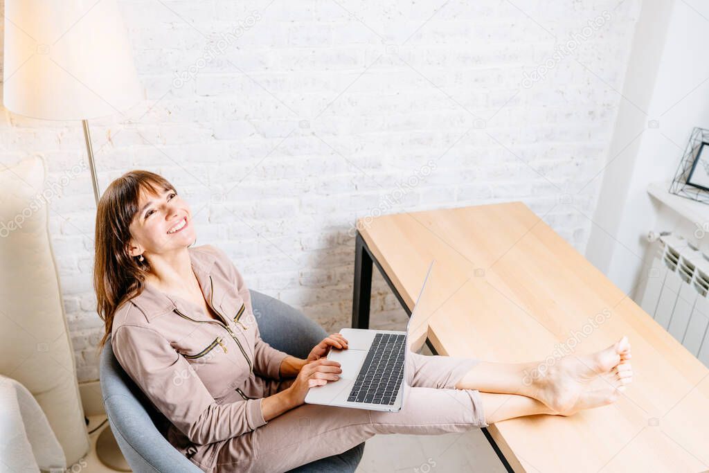 Happy beautiful businesswoman freelancer working on laptop in bright modern office at home interor. Relaxing, enjoying, smiling. Her legs on table. Comfortable concept.