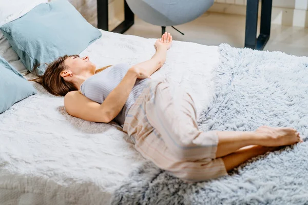 集中的なブルネットの女性は彼女を後ろに伸ばし ベッドルームでベッドの上に横たわっている側面の足と腕に目を向けました 怠惰な週末の朝 — ストック写真