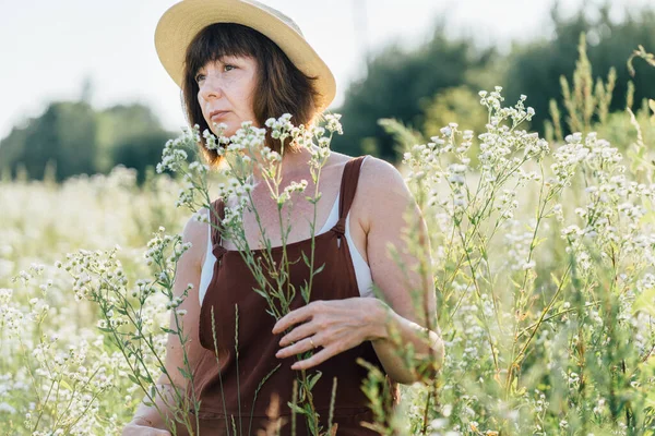 A woman in the period of menopause. Brown haired mature woman in straw hat against the field with daisies and grass background.