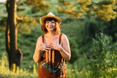 Hasır şapkalı olgun bir kadın doğada dinleniyor. Kadın bahçıvan tulumlu, ceplerinde papatya çiçekleri olan, dinlenen, bitkiler için aletler tutan. Atmosferik an, pozitif, haftasonu konsepti.