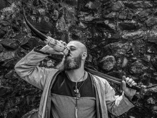 Viking warrior with thick beard and big sword drinks from the horn in front of a stone wall, black and white historical reenactment image